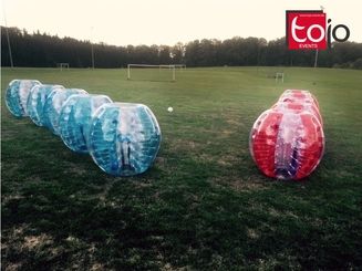 Auf einem Fußballfeld stehen mehrere transparente Bubble-Soccer-Anzüge in Blau und Rot einander gegenüber. Ein weißer Ball liegt in der Mitte. Im Hintergrund sind Fußballtore und Bäume zu sehen.