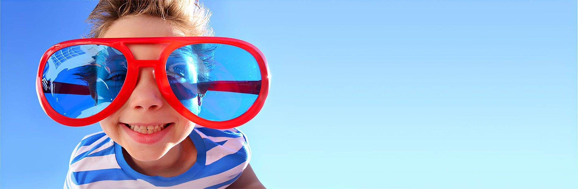 Fröhliches Kind mit blauer und weißer gestreifter Kleidung trägt übergroße rote Sonnenbrille mit blauen Gläsern und lächelt in die Kamera vor einem sonnigen, blauen Himmel.