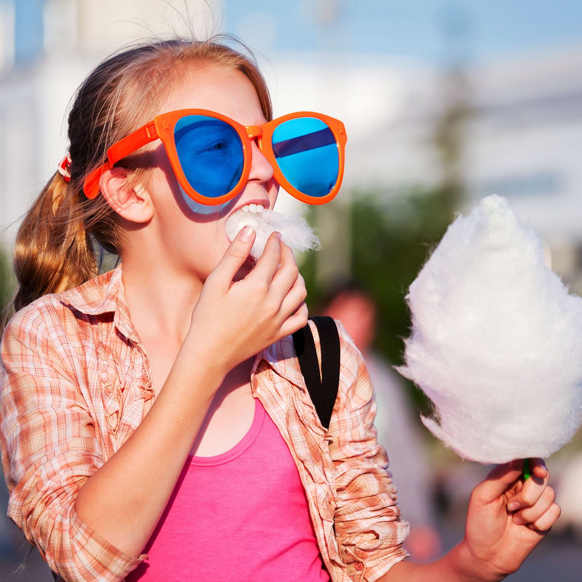 Ein junges Mädchen mit langen blonden Haaren trägt eine orangefarbene Sonnenbrille mit blauen Gläsern und isst Zuckerwatte. Sie trägt ein pinkes Oberteil und ein kariertes Hemd, während sie in einer sonnigen Außenumgebung steht.